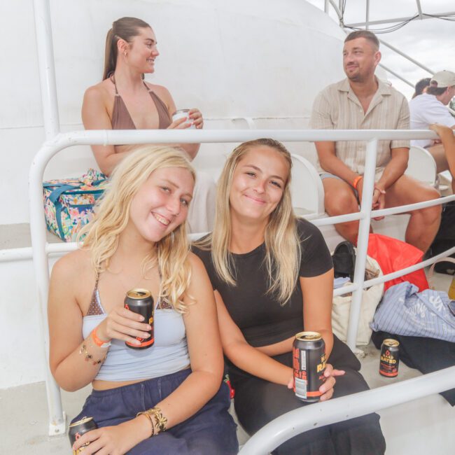 Two young women sit closely, smiling and holding canned drinks on a boat. Other people sit and chat in the background. Beach bags and belongings are beside them. The setting appears relaxed and social outdoors.