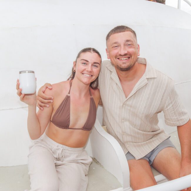 A smiling woman in a brown bikini top and beige pants sits next to a smiling man in a light shirt and shorts on a boat. The woman holds a white tumbler, and they have their arms around each other.