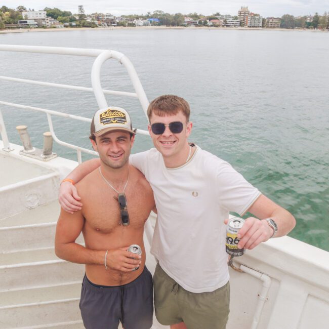 Two young men stand on a boat deck by the water, smiling at the camera. One is shirtless and wearing a cap; the other is in a white T-shirt and sunglasses. Both hold drinks, with buildings and trees visible in the background.