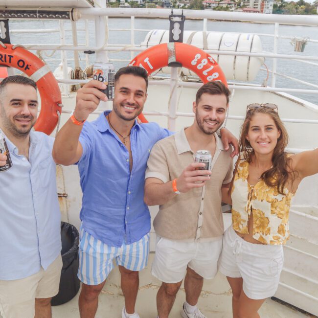 Four people, three men and one woman, are smiling and holding drinks while standing on a boat deck. Two orange lifebuoys and water are visible in the background. The group appears to be celebrating or enjoying a social event.