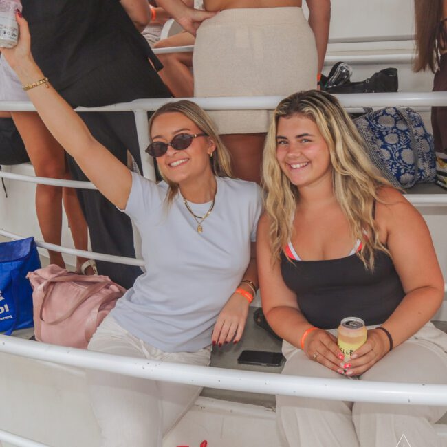 Two women sit on steps, smiling at the camera. One wears sunglasses and raises a drink, while the other holds a can. Both have wristbands and casual outfits. Other people stand around them on a white yacht.