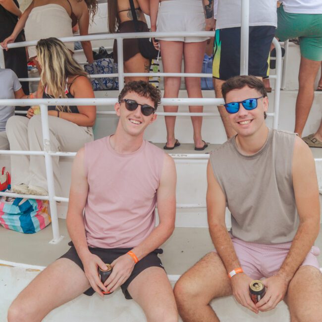 Two young men sit on the steps of a boat, both smiling and wearing sunglasses and casual summer clothes. Other people are standing or sitting in the background, suggesting a relaxed, social atmosphere.