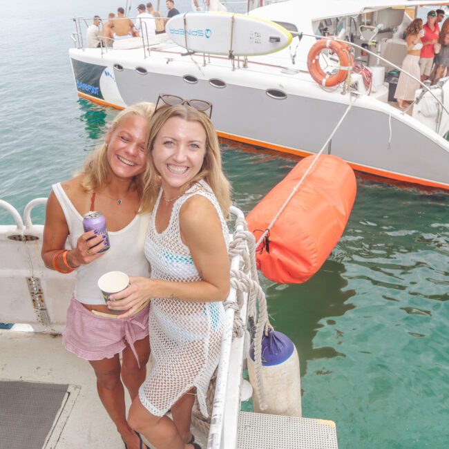 Two smiling women stand on a boat holding drinks, with a catamaran and people in the background. The water is clear, and paddleboards are visible on the catamaran. The atmosphere is bright and festive.