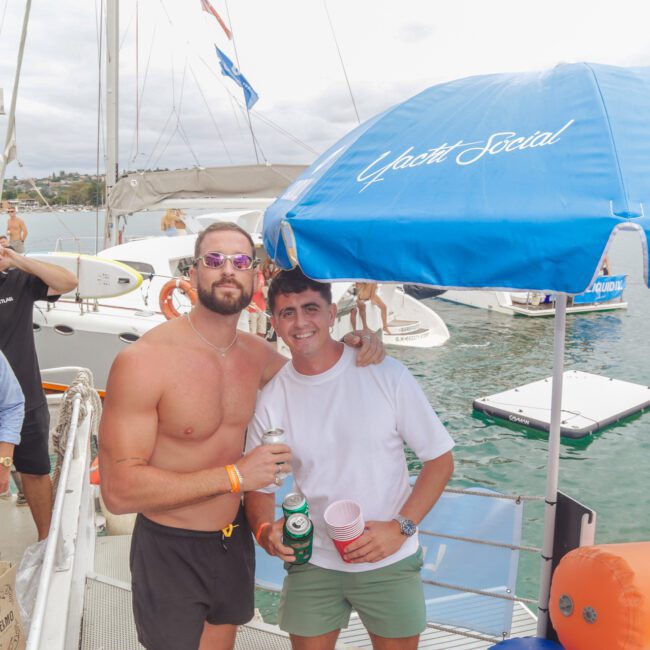 A group of young men are smiling and socializing on a yacht. Some hold drinks, while boats and water are visible in the background. A blue “Yacht Social” umbrella is open nearby, creating a festive atmosphere.