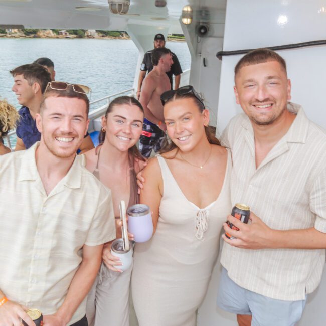 A group of four smiling people pose together on a boat, holding drinks. Other guests are visible mingling in the background, with water and coastline seen outside. The mood is relaxed and festive.