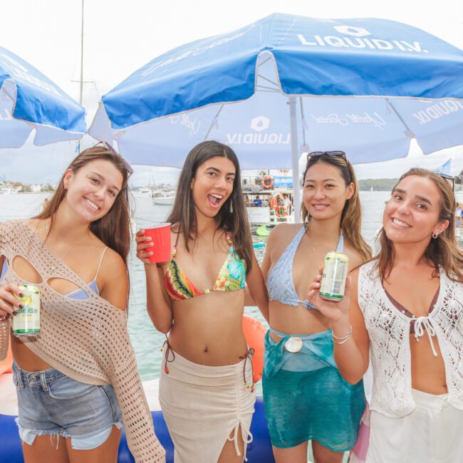 Four young women in swimsuits smile and pose for a photo at a dockside party, holding drinks, with blue umbrellas and boats visible on the water in the background.