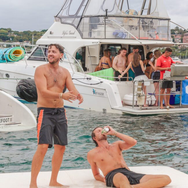 Two men are on a dock by the water, one standing and laughing, the other sitting and drinking from a can. Behind them, people are socializing on boats, with a banner that reads "LIQUID" visible. It appears to be a lively party.