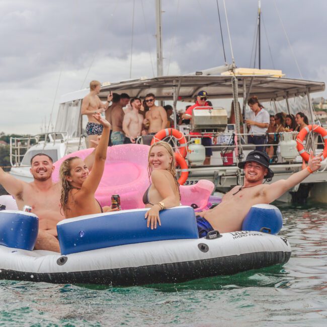 Four smiling young adults sit on a floating lounge in the water, raising drinks and arms in celebration. A lively group is gathered on a catamaran behind them under a cloudy sky, with city buildings in the background.