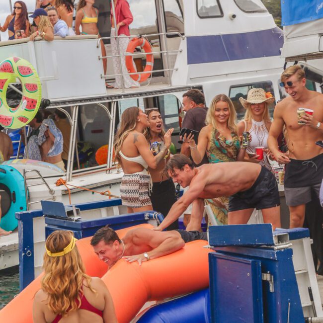 A group of people in swimsuits enjoy a boat party; some cheer as two men climb onto an orange inflatable from the water, while others watch from a nearby yacht decorated with pool floats.