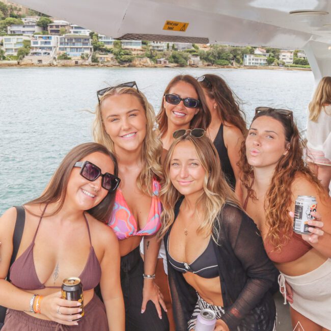 Five women in swimsuits and sunglasses smile and pose together on a boat, holding canned drinks. Houses and trees are visible across the water in the background. The atmosphere is bright and cheerful.