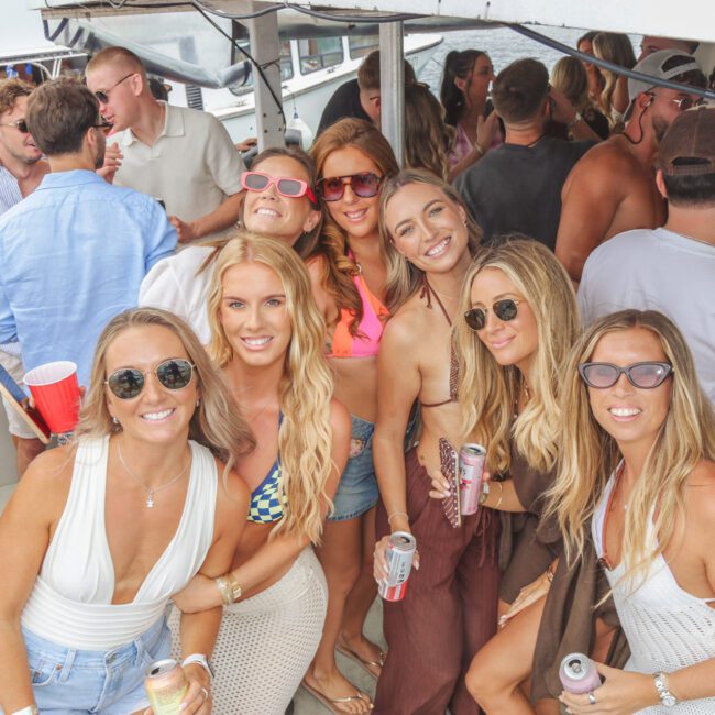 A group of smiling young adults, mostly women in swimsuits and sunglasses, gather on a crowded boat holding drinks, enjoying a lively party atmosphere.