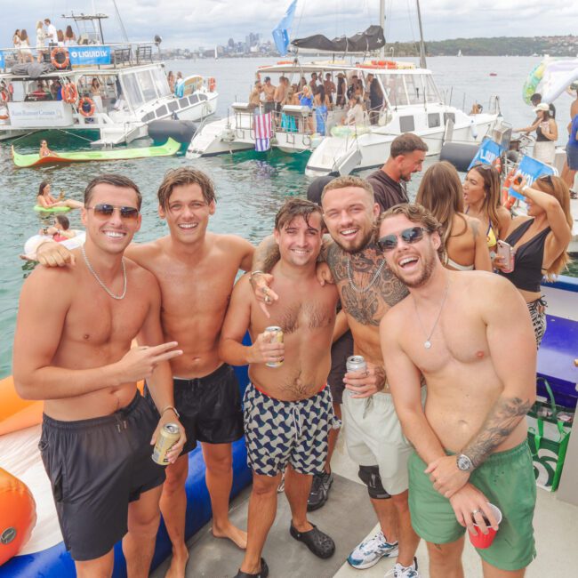 Five smiling men in swim trunks pose together at a lively boat party, holding drinks. Other people relax on boats and floaties in the water nearby, with food and city skyline visible in the background.
