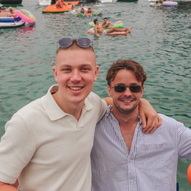 Two smiling men with arms around each other stand in front of a lake filled with people on colorful inflatables. Both wear sunglasses; one wears a white shirt, the other a blue striped shirt. The scene is lively and summery.