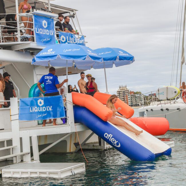 A woman slides down an inflatable slide into the ocean from a boat, while people watch and relax on the deck under blue Liquid I.V. umbrellas. The boat is anchored near other boats on a cloudy day.