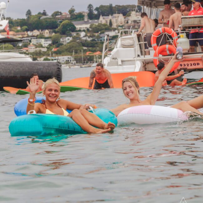 Two women in bikinis relax on inflatable rings in the water, smiling and waving. A group of people socialize on a nearby boat. The scene is festive, with overcast skies and waterfront houses in the background.