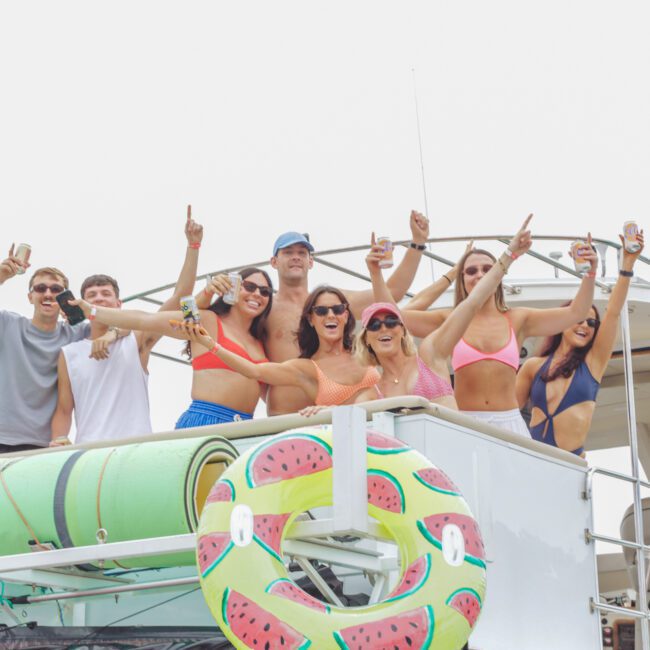 A group of young adults in swimwear smile and pose on the upper deck of a boat, holding drinks. A watermelon-patterned float is attached to the boat’s railing, and the group appears to be enjoying a sunny day.