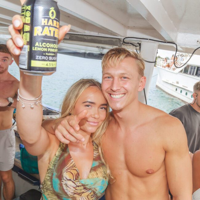 A smiling woman and man pose on a boat; the woman holds up a can and points at the camera. Other people are around them, and another boat is visible in the water outside.