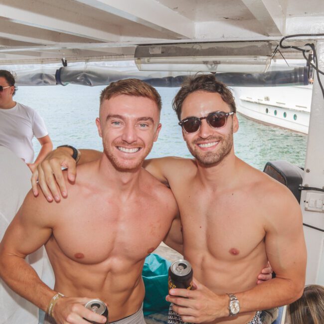 Two smiling shirtless men with drinks pose arm-in-arm on a boat. Other people are around them, some holding cups. The boat is on the water and another boat is visible in the background. It looks like a casual party scene.