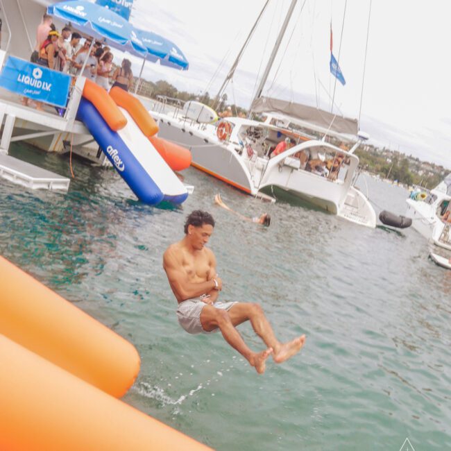 A man in swim trunks jumps off an inflatable water slide into the ocean near several anchored yachts, with people relaxing on deck under blue umbrellas.