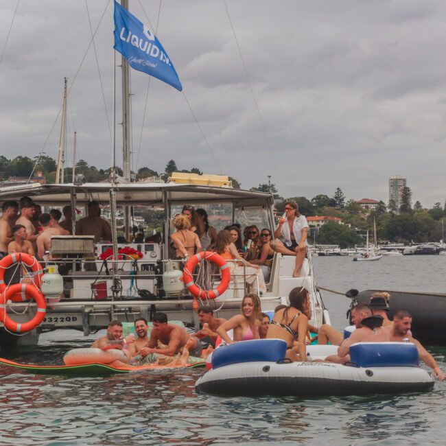A group of people relax and socialize on a catamaran and inflatable floats in the water near a coastal city, under a cloudy sky. Some are on the boat and others are enjoying the water and floats nearby.