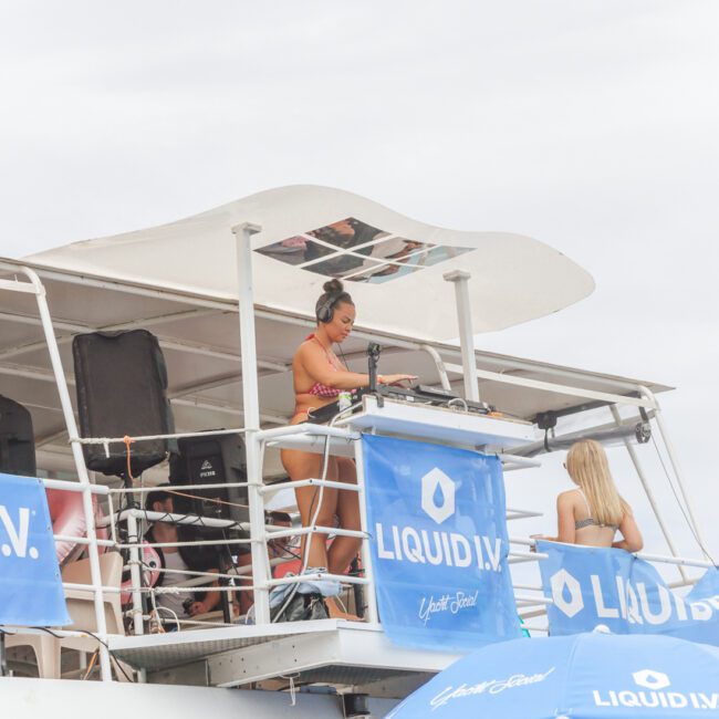 A woman in a bikini is DJing on the upper deck of a boat decorated with blue Liquid I.V. banners, surrounded by people enjoying a party under a cloudy sky.