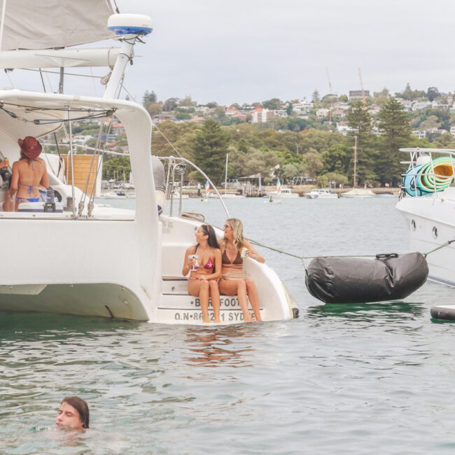 Two women in swimsuits sit on the back of a yacht, smiling and talking. Another person is swimming nearby in the water. Trees, houses, and other boats are visible in the background under a cloudy sky.