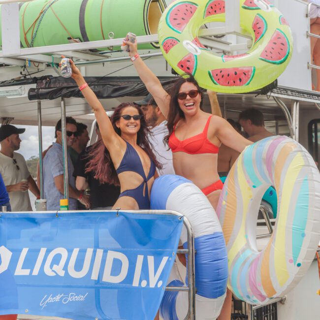 Two women in swimsuits smile and raise their arms on a boat decorated with colorful pool floats. Other people are on the boat, enjoying drinks. A "Liquid I.V." banner is visible on the railing.
