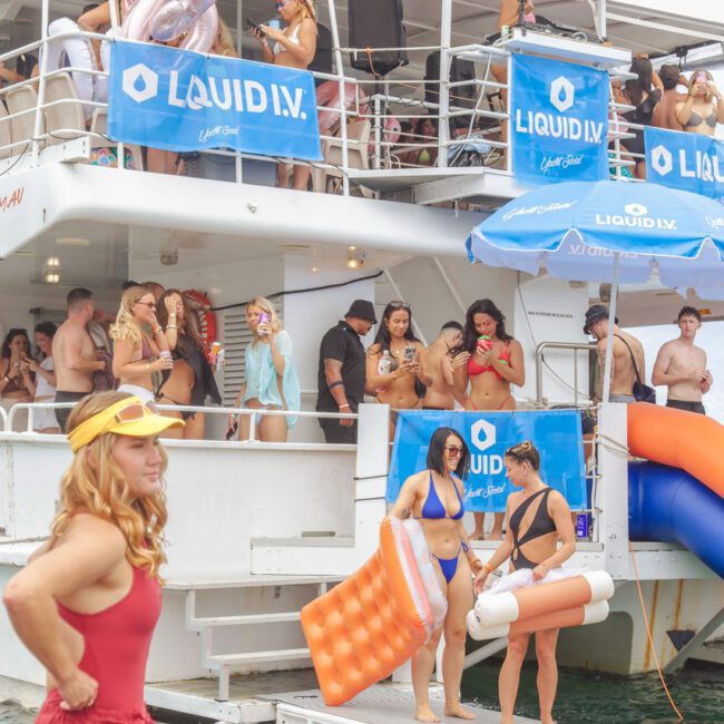 A lively boat party with people in swimwear on and around a yacht, holding inflatables. A woman in a red lifeguard outfit stands in the foreground. Blue "Liquid I.V." banners decorate the boat.