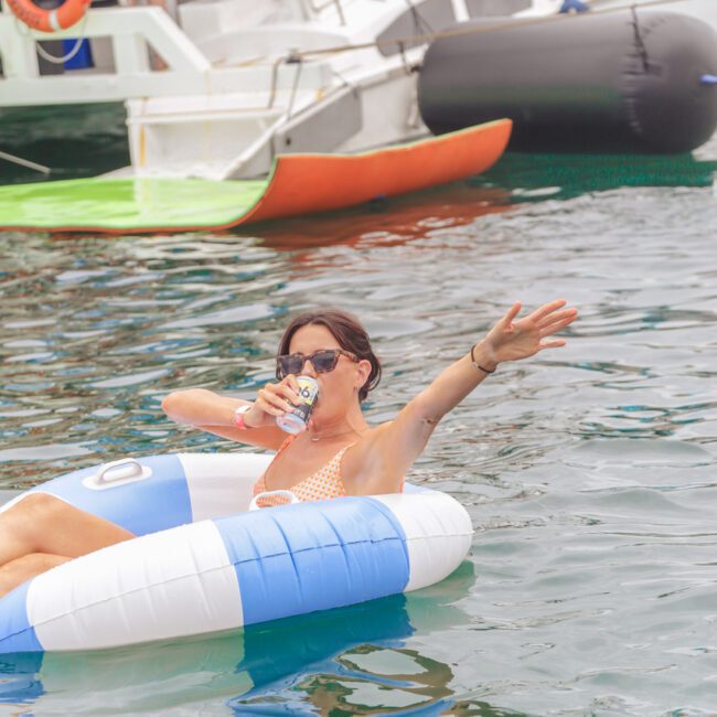 A woman in sunglasses and a swimsuit relaxes on a blue and white inflatable ring in the water, holding a drink and raising one arm. Boats and people are visible in the background.