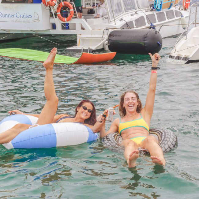 Two women in swimsuits relax on inflatable tubes in the water, smiling and raising their arms joyfully. Boats labeled "Rum Runner Cruises" are docked behind them. The scene is bright and cheerful.