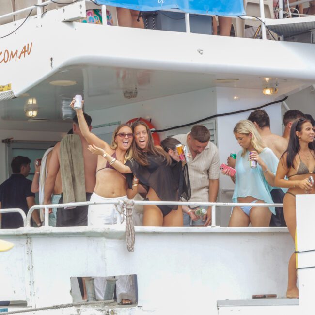 A group of young adults in swimsuits enjoy a lively party on a boat, holding drinks, laughing, and dancing near the railing. Banners and party decorations are visible in the background.