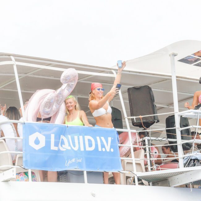 A group of people on a yacht party stand on the upper deck. One woman in sunglasses and a pink bandana raises a blue cup. A blue banner with “LIQUID I.V.” hangs on the railing. The mood is festive and bright.