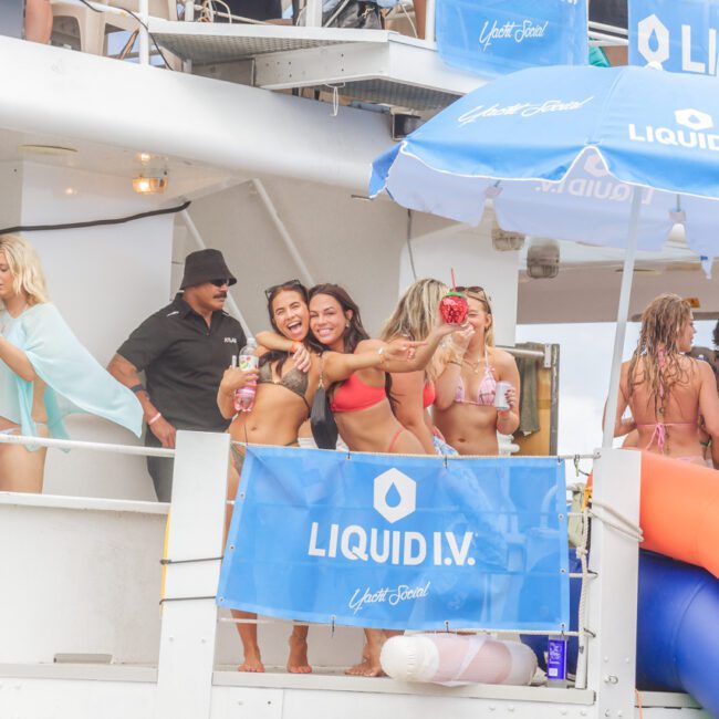 A group of women in swimsuits pose and smile on a boat deck decorated with "Liquid I.V." banners, while others stand nearby. The scene is lively and summery, with an umbrella and drinks visible.