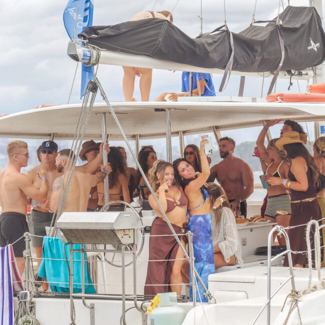 A group of people in swimwear are gathered on a yacht, socializing and taking selfies. Some are standing or sitting under the shaded deck, while others are on top. The atmosphere is lively and festive.