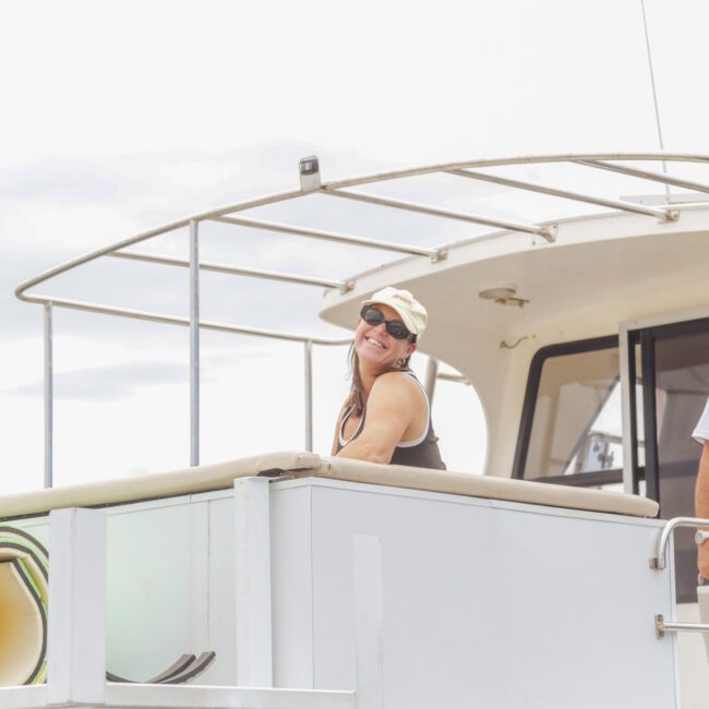 A smiling woman in sunglasses and a hat leans on the railing of a boat, while a man in a white shirt and cap stands nearby. Colorful coiled hoses are visible on the deck under a cloudy sky.