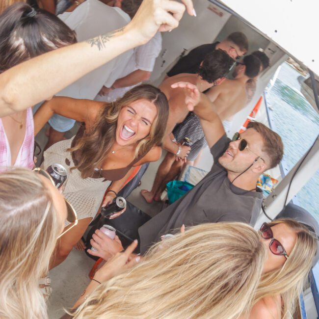 A group of people on a boat dancing and laughing, holding drinks. Everyone appears to be having fun, with one woman in the center smiling widely. The boat is on the water under a sunny sky.