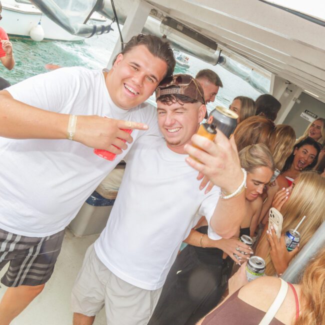 Two men smile and pose for the camera on a crowded boat, each holding drinks. Other people are socializing in the background, enjoying a lively atmosphere on the water.