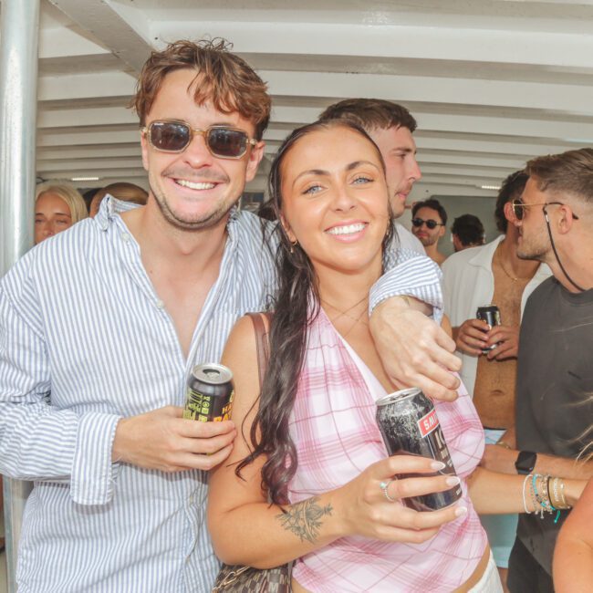 A man in sunglasses and a striped shirt stands with his arm around a smiling woman in a pink top, both holding drinks on a crowded boat with other people socializing in the background.