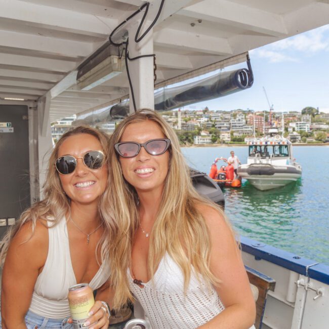 Two women in sunglasses smile at the camera on a boat; one holds a drink. In the background, another boat is on the water, and there are buildings and greenery along the shoreline under a partly cloudy sky.