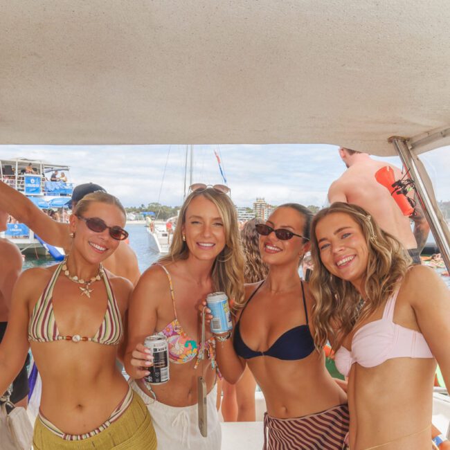 Four women in bikinis and sunglasses smile and pose together on a boat, each holding a drink. The background shows other people, boats, and water under a sunny sky. The atmosphere is festive and relaxed.