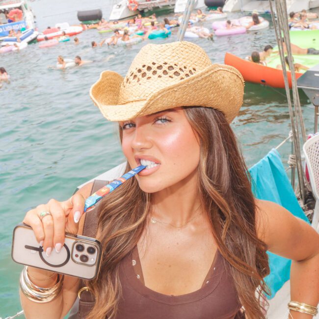 A woman wearing a straw cowboy hat poses on a boat, holding her phone and playfully biting a colorful straw. People and boats are visible on a lake in the sunny background.
