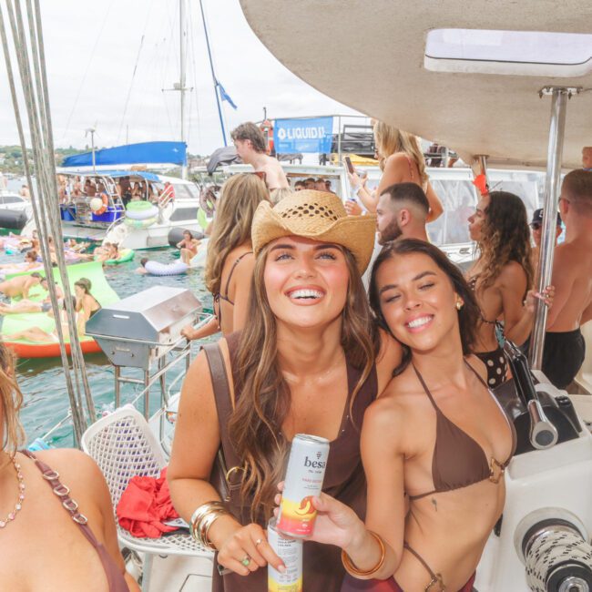 Three young women in swimsuits smiling and posing on a crowded boat during a lively summer party, with people on floaties and other boats in the background on the water. The atmosphere is festive and energetic.