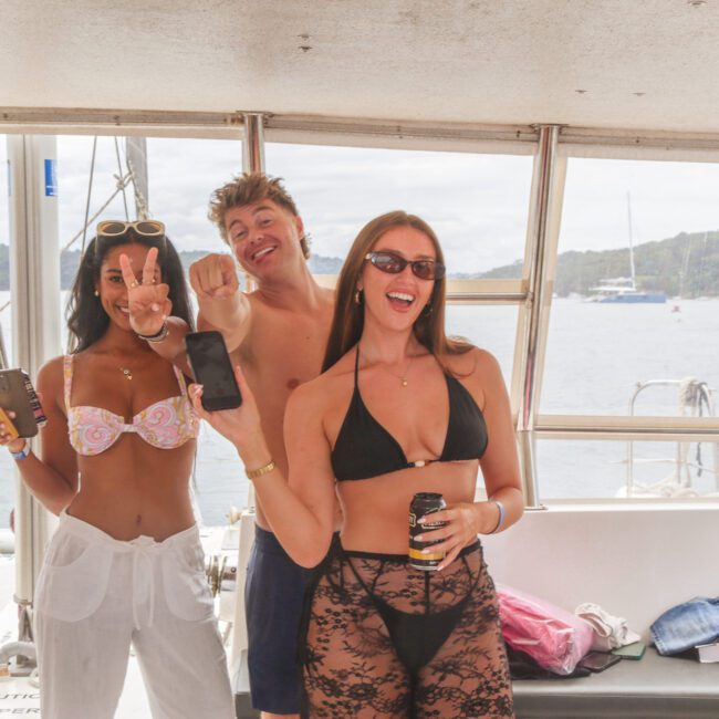 Three people in swimwear smile and pose on a boat, holding drinks and phones. The group is enjoying a sunny day on the water with a scenic background of hills and anchored boats.