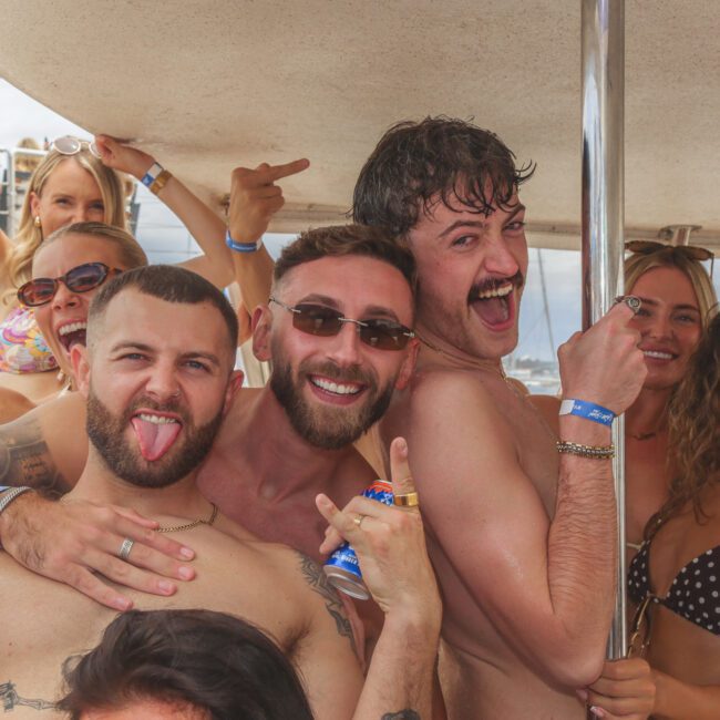 A group of smiling people in swimsuits pose together on a boat, making playful gestures and holding cans, with sunlight and water visible in the background.