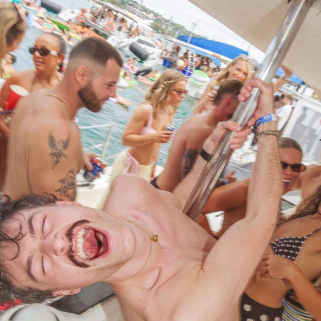 A man laughing and hanging onto a pole on a crowded boat, surrounded by people in swimsuits enjoying a lively party on the water with other boats and floats in the background.