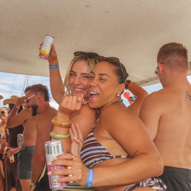 Two women in swimsuits smile and pose with canned drinks at a lively boat party, surrounded by other people enjoying the sunny day. The scene is festive and bright, with bracelets on their wrists.