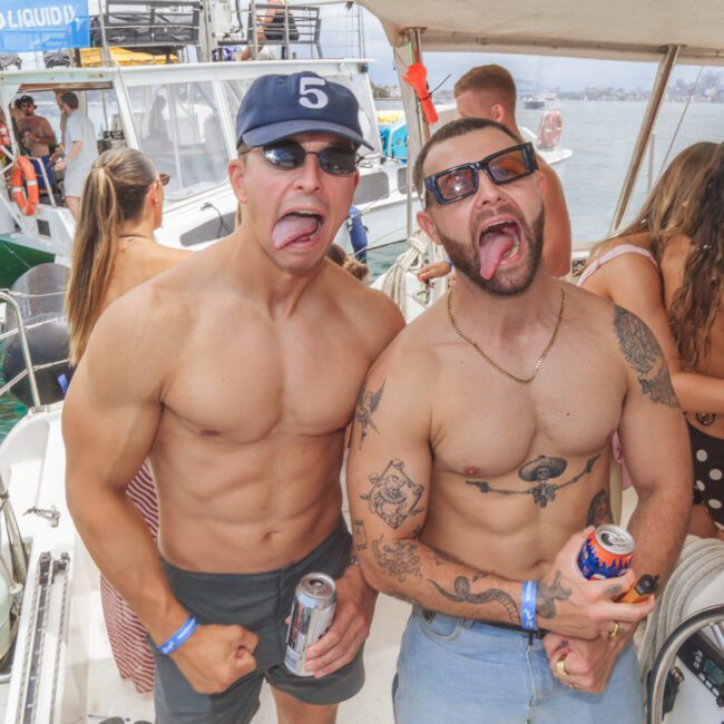 Two shirtless men wearing sunglasses and making funny faces pose on a boat with drinks in hand. Other people are mingling in the background, and nearby boats float on the water under a sunny sky.