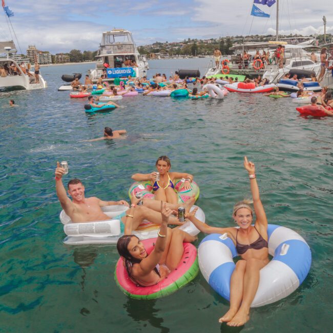 A group of people relax on colorful inflatable pool floats in the water, holding drinks, with boats and more people on floats in the background at a lively outdoor party.