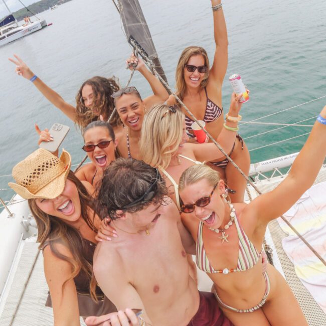 A group of young adults in swimsuits joyfully pose on a sailboat, smiling and holding drinks, with the ocean and another boat visible in the background. One woman wears a cowboy hat.