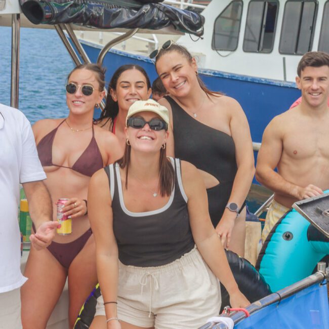 Five people in swimwear smile and pose together on a boat, with diving and swimming gear around them. A large boat and water are visible in the background. It appears to be a sunny, fun day out on the water.